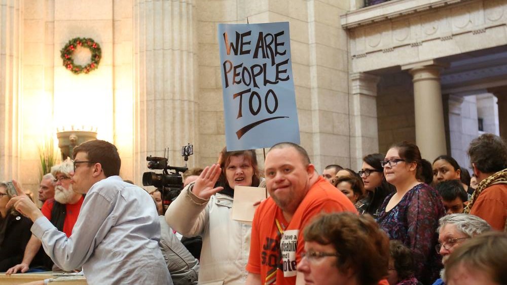 People with disabilities wave a sign saying, 'We are People Too,' while advocating for themselves at the Manitoba Legislature. 